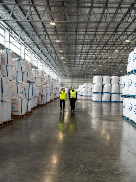Workers walking inside a large warehouse construction project with high ceilings and stacked bulk storage bags.